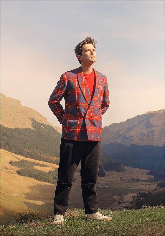 Man in a red and blue plaid blazer and black pants standing on a hill with mountainous terrain in the background, looking upwards with a serene expression.