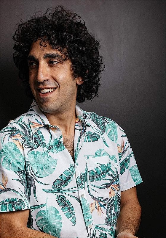 Portrait of a smiling man with curly hair looking to the side, wearing a white tropical print shirt against a dark background.