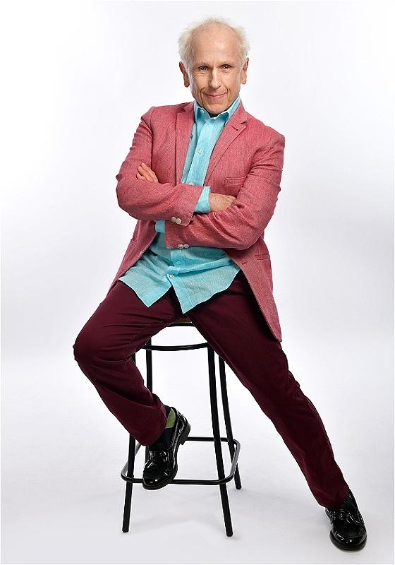 Elderly man with white hair posing confidently on a stool, wearing a coral blazer, turquoise shirt, and maroon trousers.