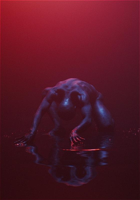 A dark figure crouching in water, illuminated by a red light with a reflective surface creating a mirror image below.