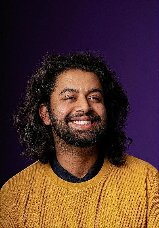 Portrait of a joyful man with long curly hair, wearing a yellow sweater over a blue shirt, smiling broadly against a purple background.