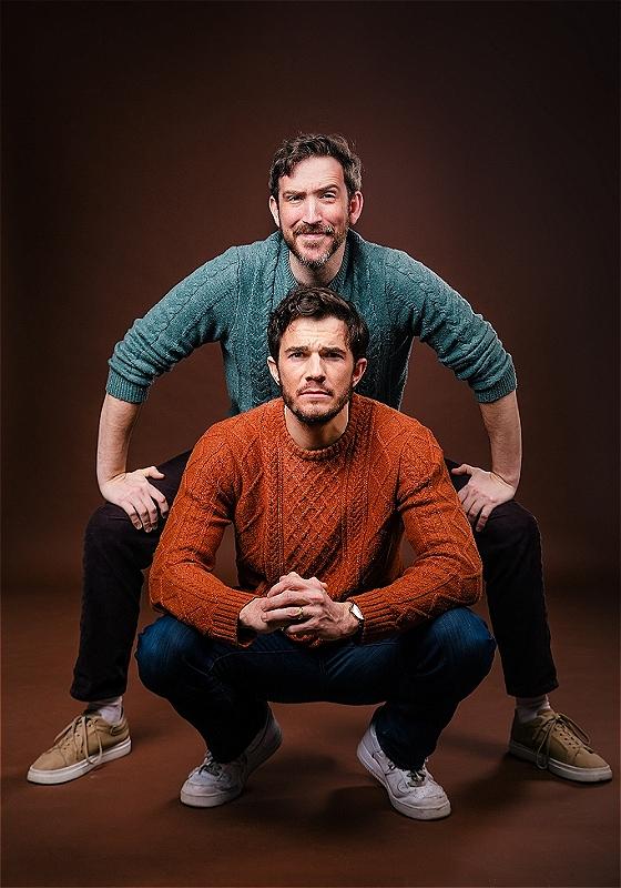 Two men posing in a studio, with one crouching far down and the other straddling behind him, both wearing knitted sweaters and jeans, set against a brown backdrop. The crouched man wears an orange sweater and white sneakers, while the straddling man wears a green sweater and beige sneakers, both smiling slightly at the camera.