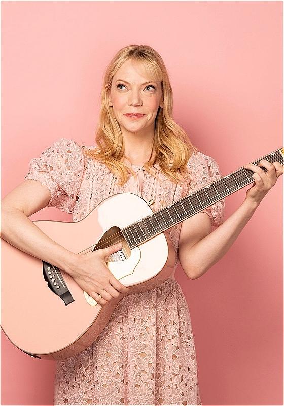 Woman with blonde hair in a pink lace dress smiling and playing a pink acoustic guitar against a pink background.