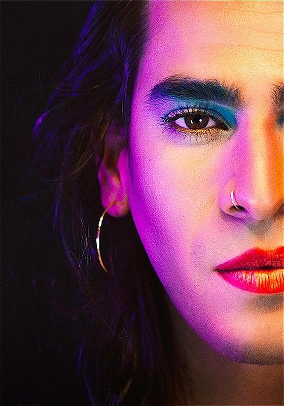 Close-up portrait of a person with long wavy hair, dramatic blue eyeshadow, a nose ring, and red lipstick. The lighting casts vivid pink and blue hues across their face.