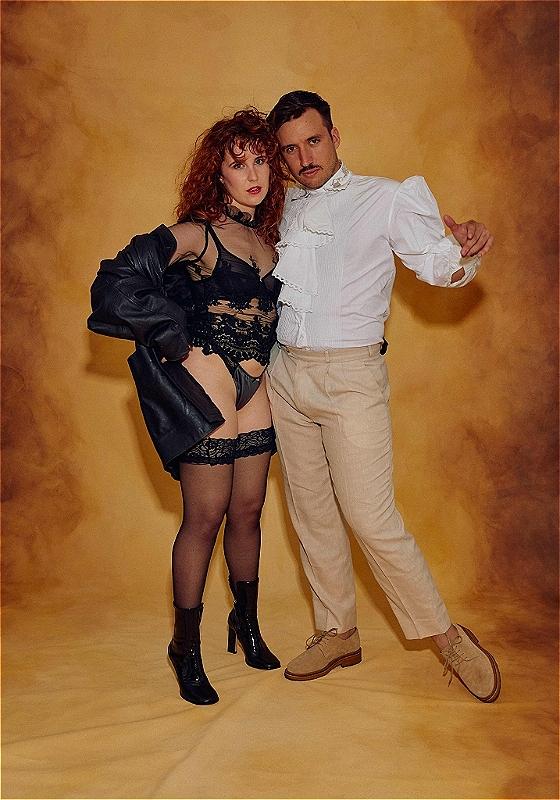 Two people posing dramatically against a golden yellow backdrop, with the woman wearing a black lace lingerie set and a leather jacket, and the man in a high-collared white shirt and beige trousers. Both are looking intensely at the camera.
