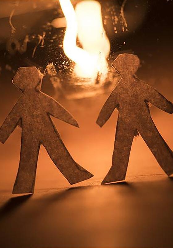 Two paper cutout figures of people walking toward each other with a large flame in the background, all immersed in a warm, amber light.