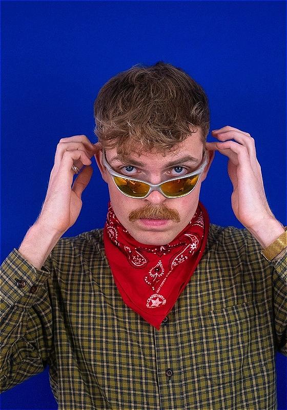 In front of a blue background Horatio looks directly at the camera. He is wearing a green checkered shirt, a red bandana around his neck.