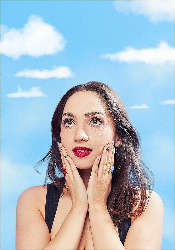Young woman with long dark hair looking up in surprise or wonder, hands on cheeks, wearing a black sleeveless top against a cloudy blue sky background. Her nails are long with multi-colored polish, and she has vibrant red lipstick.