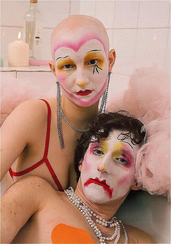 Two people pose in a bathtub with dramatic clown-style makeup. Around the bath there is pink tulle and a candle is lit.