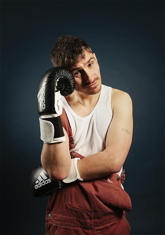 Young man with a contemplative expression wearing boxing gloves and a white tank top, leaning his cheek on one glove against a blue background.
