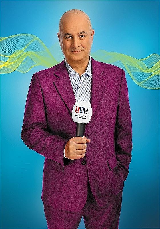 Iain Dale in a purple suit jacket and light blue shirt , holding a microphone labeled 'LBC', standing in front of a blue background with abstract yellow and green waves.