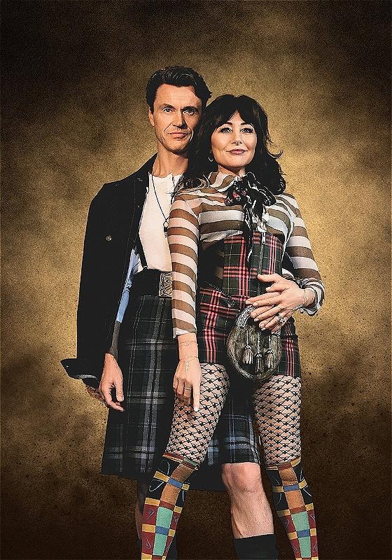 A man and a woman dressed in avant-garde Scottish-inspired outfits including tartan kilts, patterned fishnet stockings, and colorful mismatched socks, standing closely together against a textured golden background.