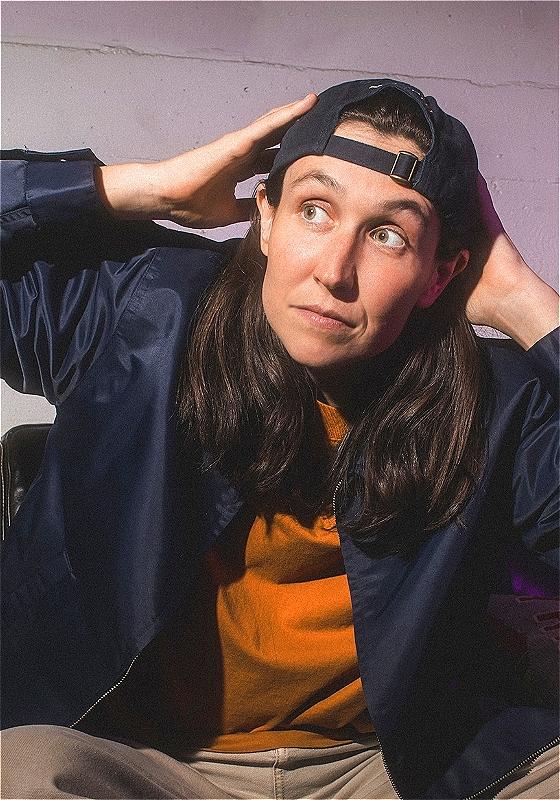 A young woman with long brown hair wearing a backwards baseball cap, navy blue bomber jacket, and orange t-shirt, sitting with a playful expression and hand on her head against a light purple background.