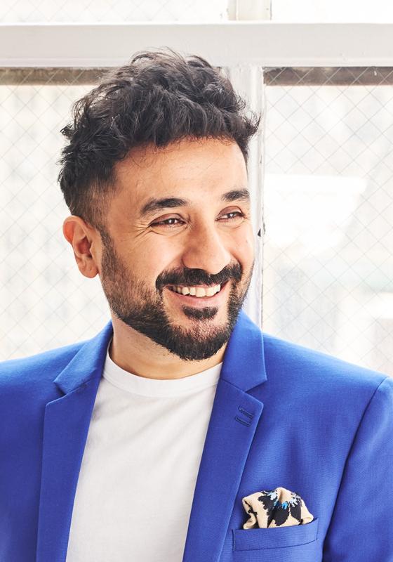 Vir Das looks smilingly off camera and wears a blue suit with a white shirt.