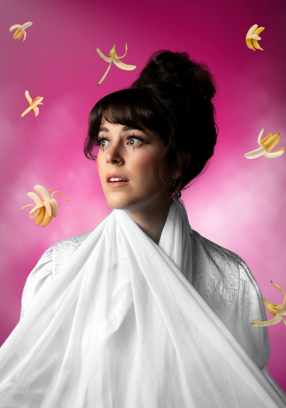 Woman with dark hair up in a bun, wearing a white scarf, gazing upward against a pink background with floating yellow banana shapes.