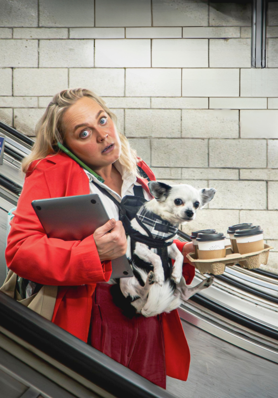 A busy looking woman in a red suit and corporate wear stands on an escalator holding a laptop, dog in a harness and four takeaway coffee cups in a tray.