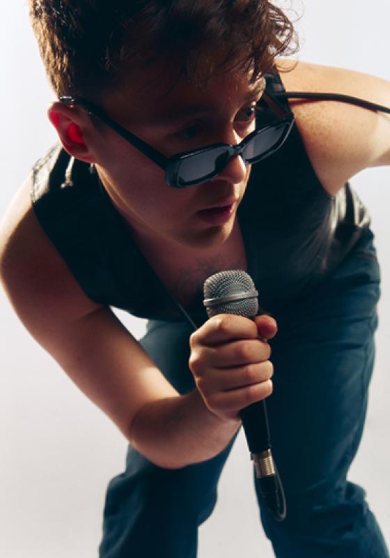 Jake Roche, arms bare in a waistcoat, jeans, and stylish sunglasses, crouches with a microphone in hand. Jake is lit from the side, and the background is white. 