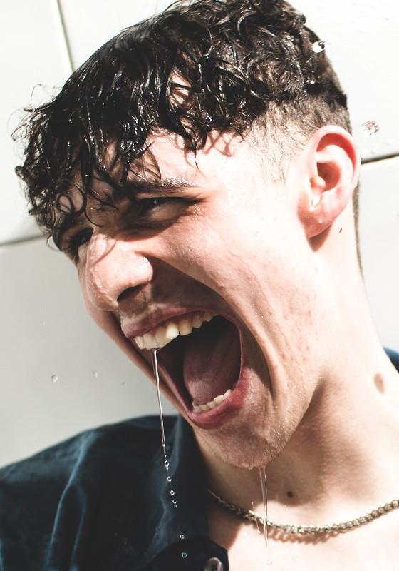 Young man with wet curly hair, wearing a dark shirt, laughing or shouting with joy as water drips from his mouth, against a sunlit white tiled wall.