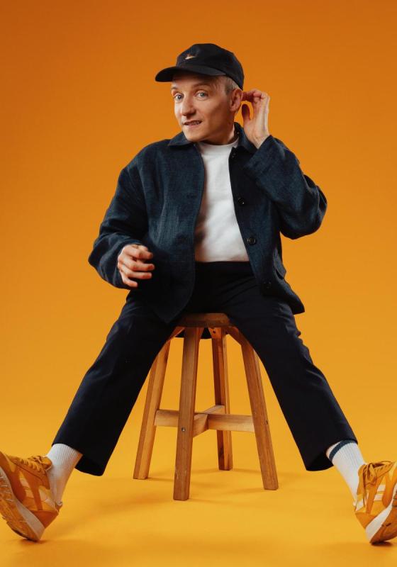 Will sits on a kitchen stool, legs outstretched with his feet pointed upwards. He smiles awkwardly, touching his ear with his left hand. 