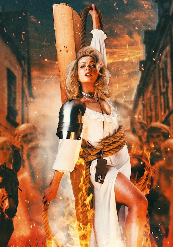 Abi wearing white shirt and shoulder armour is tied to a plank of wood with sparks of fire surrounding her. Blurred silhouettes of historical figures make up a crowd behind her.