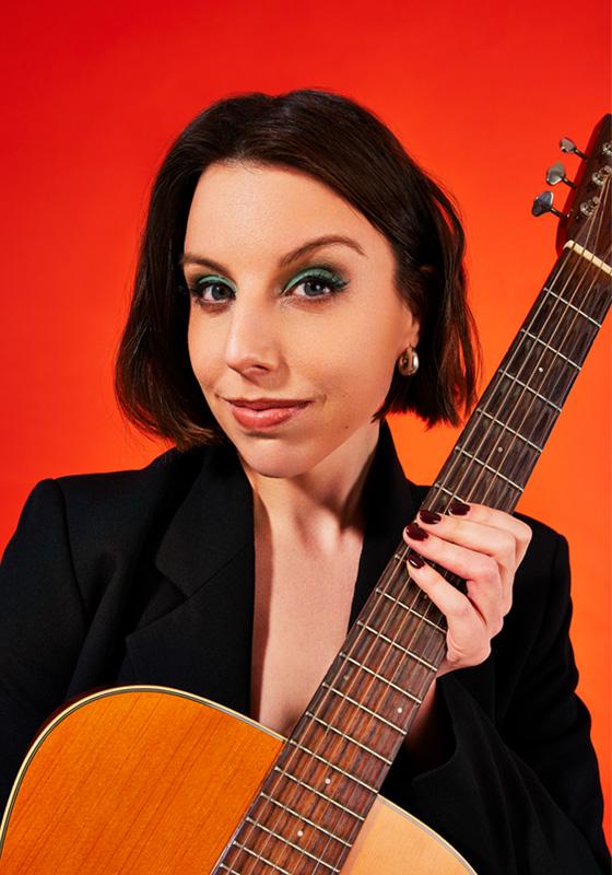 Isobel smirks at the camera holding up a guitar.