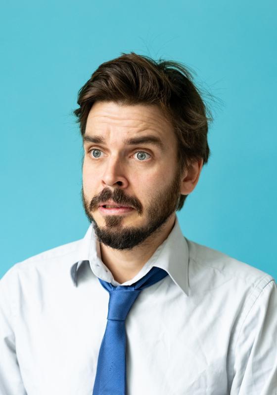 Bearded man in white shirt and blue tie looking puzzled in front of a blue backdrop.