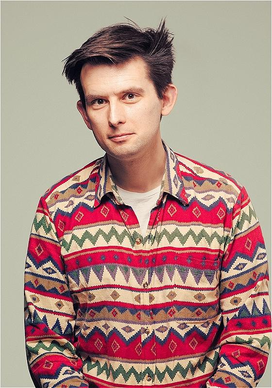Kieran in a funky printed top against a plain grey background looking at the camera