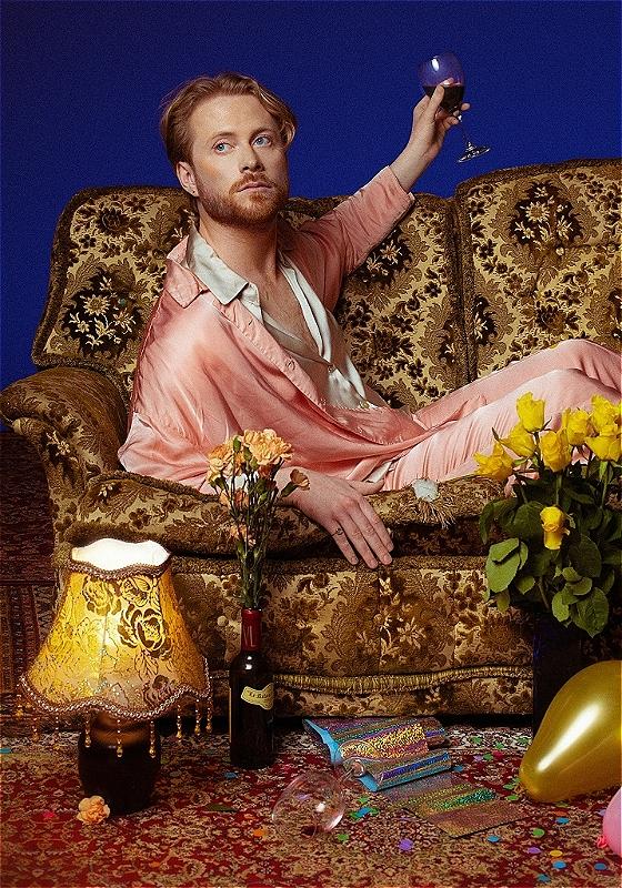 Performer sitting on gold sofa holding wine glass wearing pink silk pjs surrounded by balloons, flowers, wine and lamp