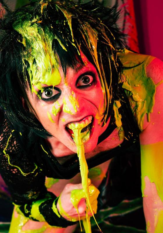 A person with black hair and dark eye makeup is covered in bright green and yellow paint, appearing animated and making an expressive face with an open mouth.