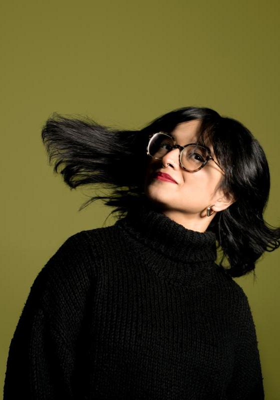 Headshot of Shalaka flipping their hair. Performer wears a black polo neck jumper and red lipstick.