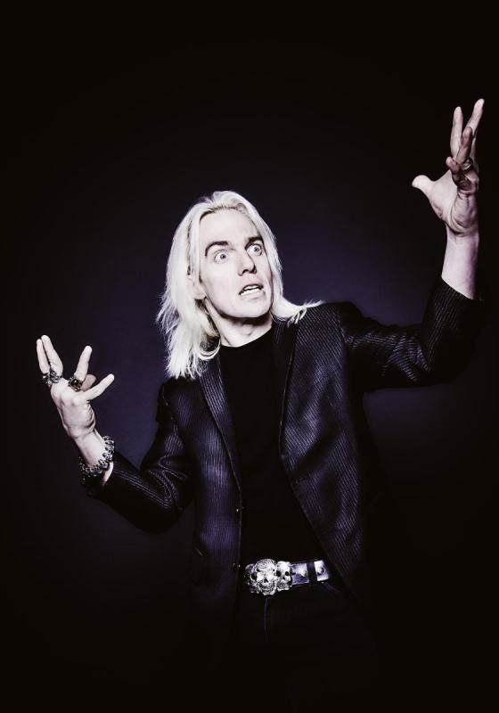 John Robertson wearing all black against a black background gesticulating wildly with a crazed look on their face