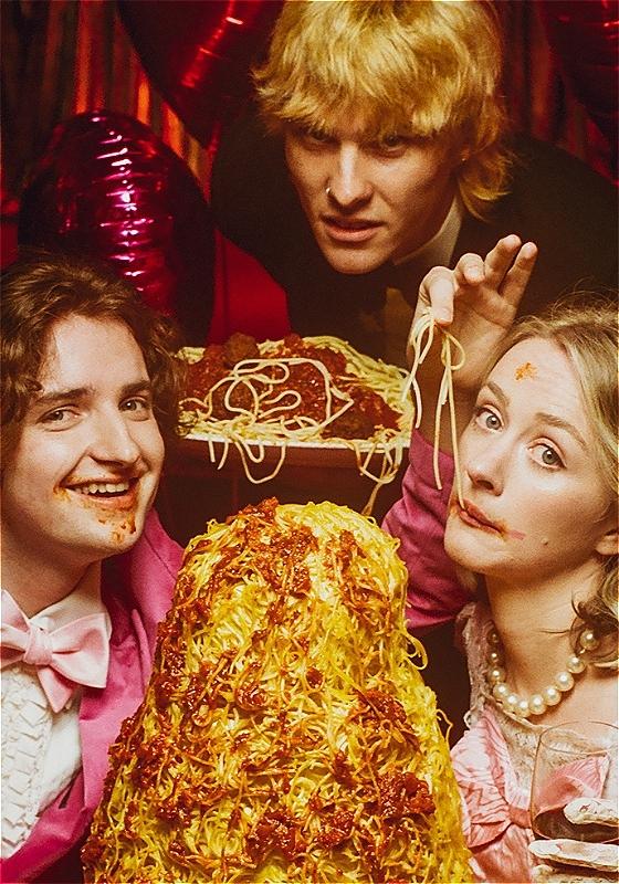 A third man stands looking garishly between a woman and a man who are eating spaghetti, Lady and the Tramp style. There is spaghetti and tomato sauce around the couples faces.There is a mountain of spaghetti in front of her. It has an 80s wedding vibe about it, a lot of pink. 