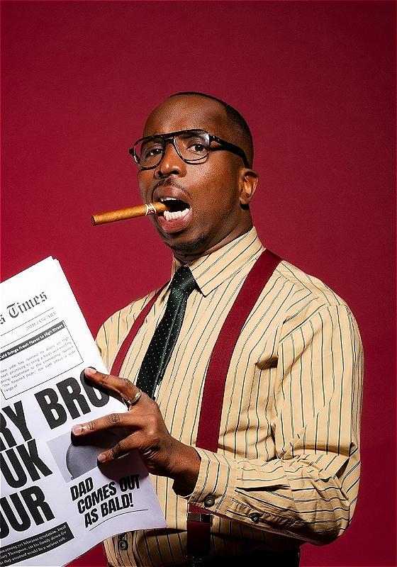 Kiell Smith-Bynoe looks to the camera with black glasses on and a cigar in his mouth. He wears a pink shirt with thin black stripes, a black tie and burgundy braces. He holds a newspaper in his hand. 