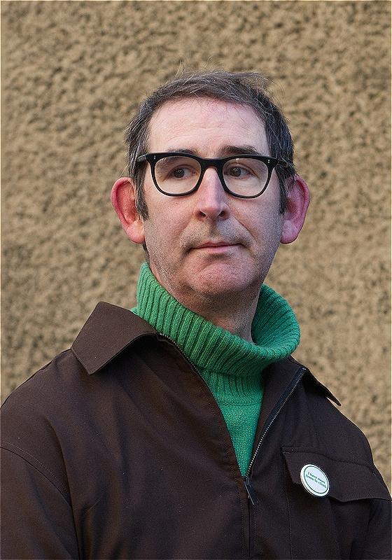 Man wearing black glasses, a green jumper and brown jacket.