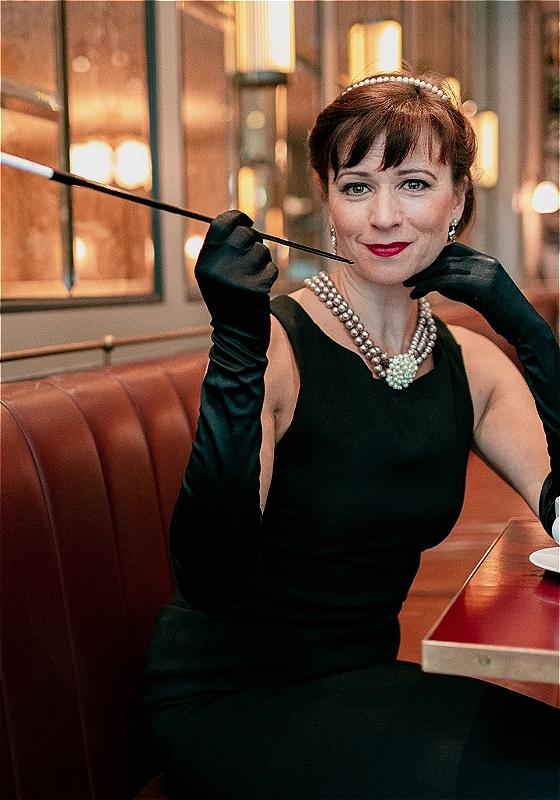 A woman wearing a black dress, gloves and pearls, sitting in a booth with a cup and saucer.