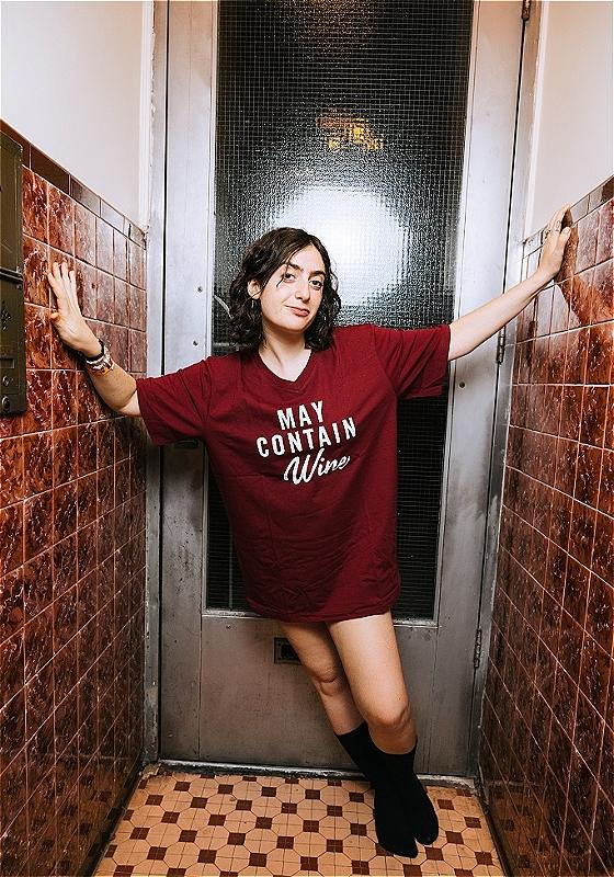 Illustration of woman standing in a door way wearing red t-shirt reading 'may contain wine'. 