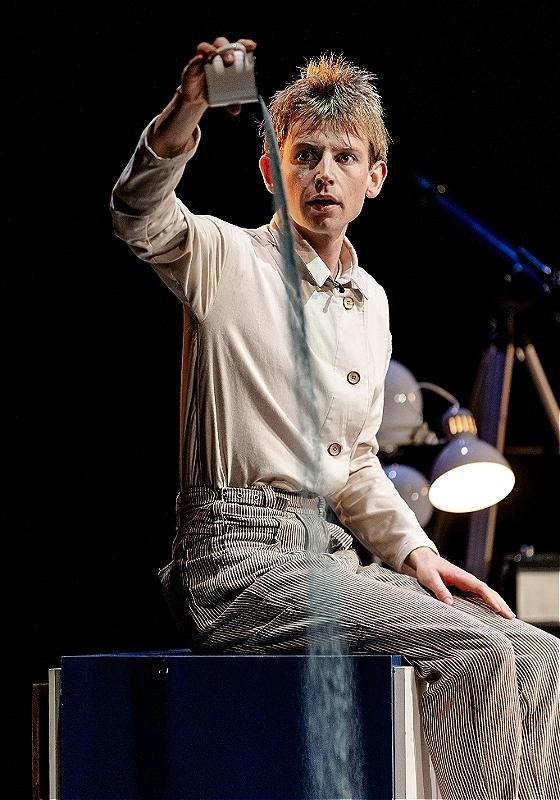 The performer pouring something out of a cup, seated next to a lamp.