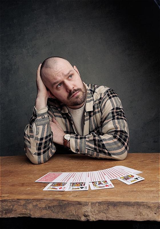 A man sitting at a desk, looking upwards. In front of him on the table are a deck of cards. 