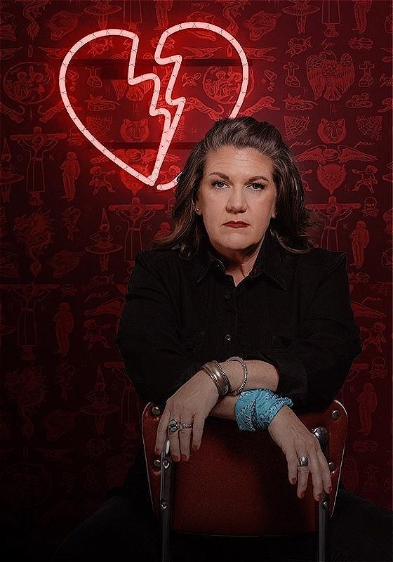 A woman sitting in front of a lit-up neon pink broken heart sign. 