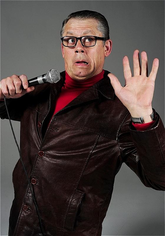 John in a brown leather jacket and red shirt holds a microphone, gesturing with one hand, against a grey background.