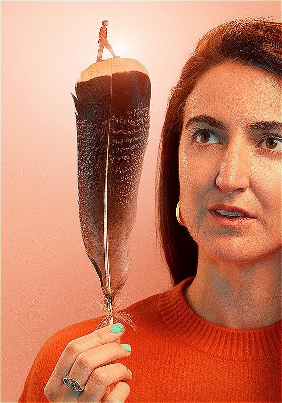 A person holding a large feather, with a miniature figure standing on top, against a soft pink background.