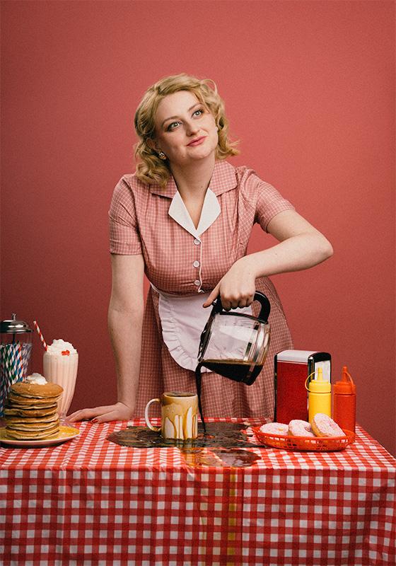 Lorna wearing a red gingham dress with an apron smiling sweetly as she pours coffee into an overflowing cup.