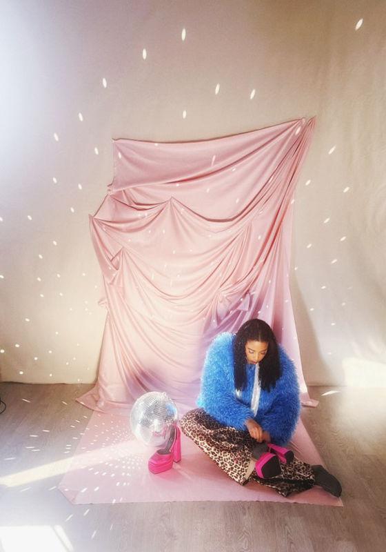 Performer sitting on the floor putting her pink platform heels on amidst a disco ball and pink fabric backdrop