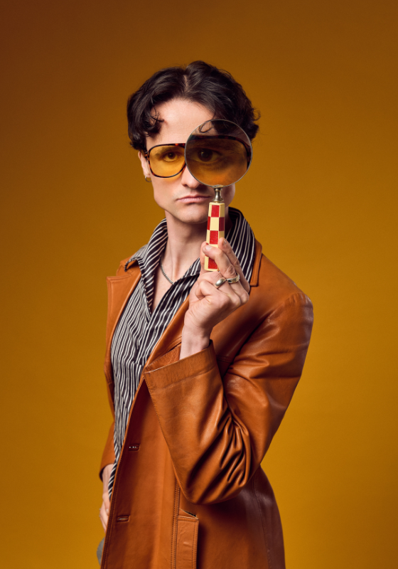 Performer holds a magnifying glass in front of his eye. He wears an orange leather coat jacket and 70s style tinged sunglasses against an orange background.