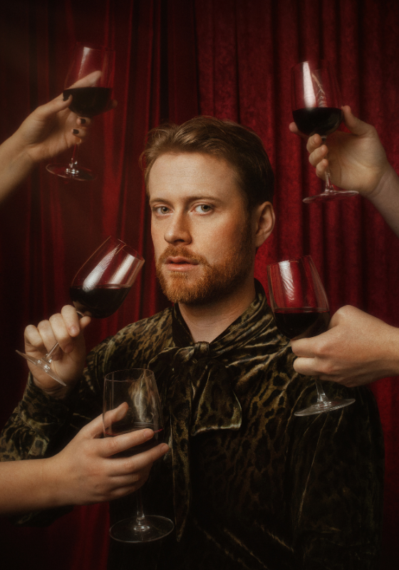 A sultry looking John is surrounded by full glasses of red wine held by outstretched hands.