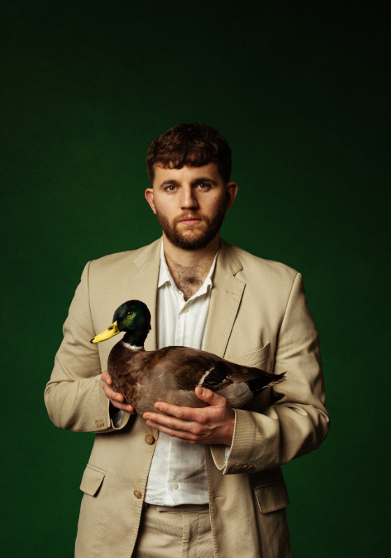Rory holds a mallard duck awkwardly wearing a beige suit against a dark green backdrop.