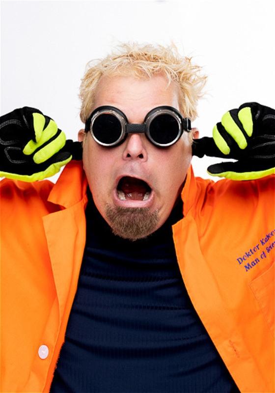 Man wearing black goggles, and gloves in an orange lab coat. Stands with his mouth agape in shock and his fingers in his ears, directly looking into the camera.