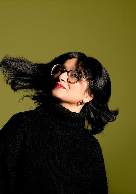 Shalaka flings her black hair, wearing a black turtleneck on a green background