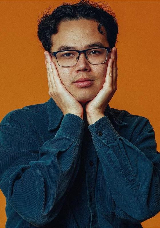 James Trickey holds his face in his hands as he wears a blue jumper against an orange background