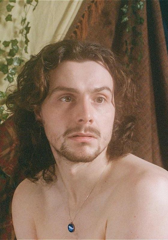 Sam, shirtless, stares into the distance behind the camera. There are vines and curtains draped in the background. 